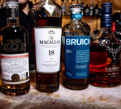 Image Of Four Rare Scotch Whisky Bottles From Glenrothes, Macallan, Bruichladdich, And Dalmore Displayed On A Table At Wevino In Westlake Village, Set In A Warm, Candlelit Ambiance.